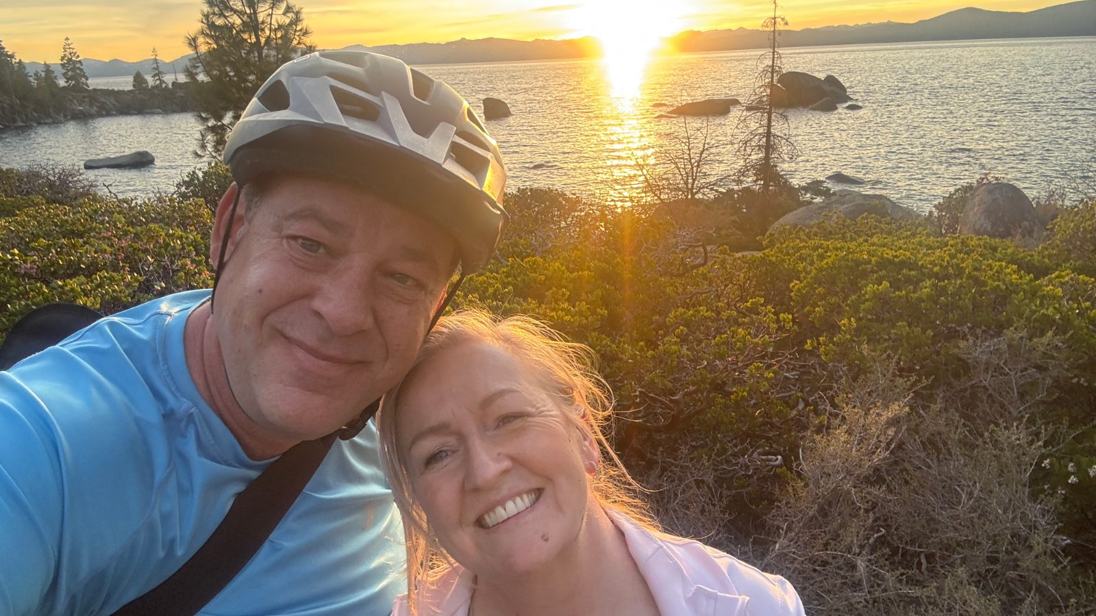 7:08 PM: a sunset selfie, Christian still in his bike helmet and Helena beside him, the sun low over the lake behind them.